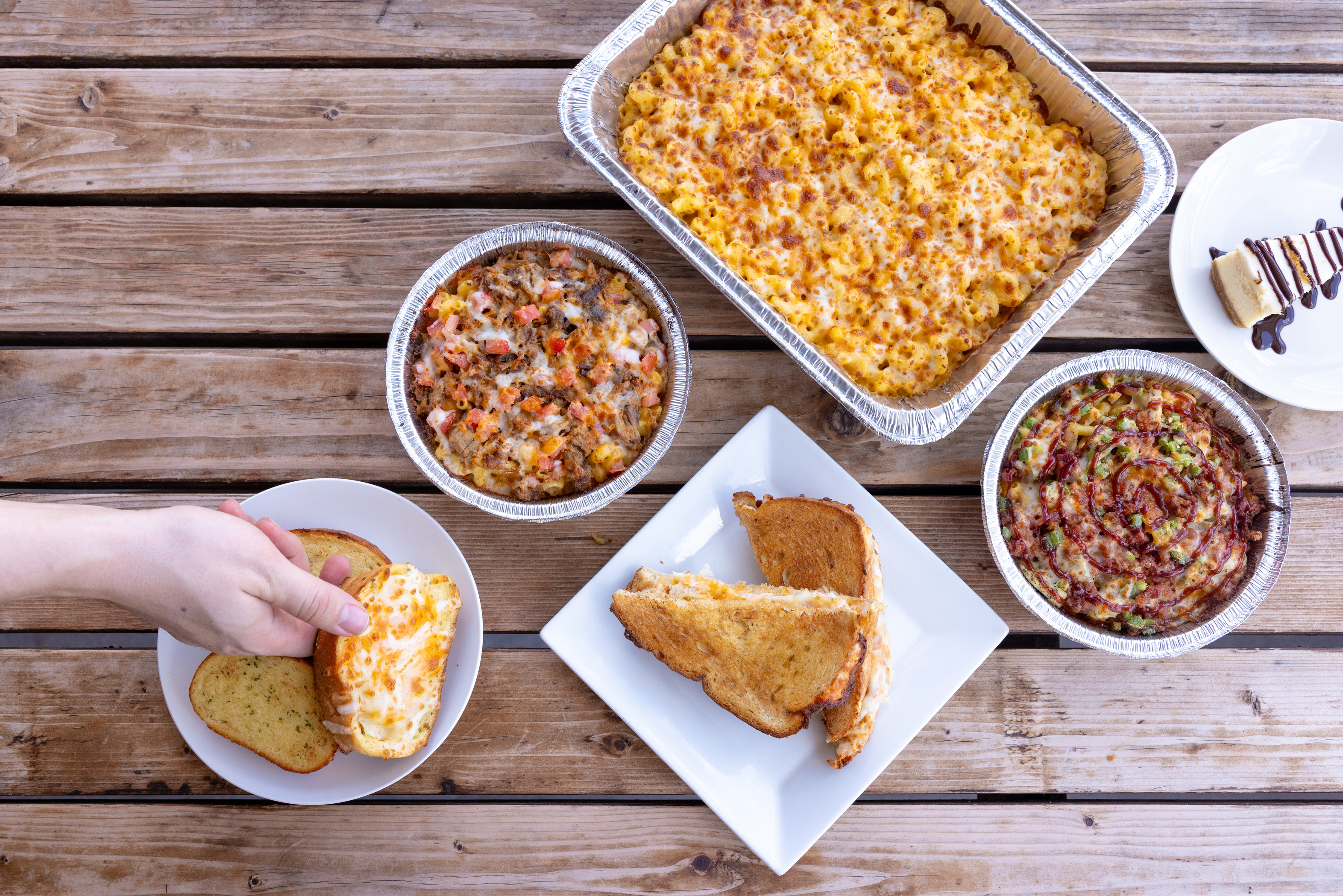 Mac and cheese party trays, mac bowls, grilled cheese, garlic bread, and cheesecake on a wooden table